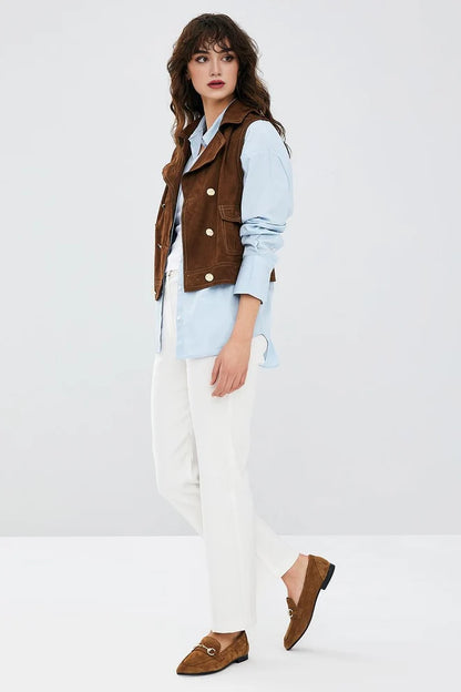Woman wearing a brown vest, light blue shirt, white pants, and brown shoes on a white background